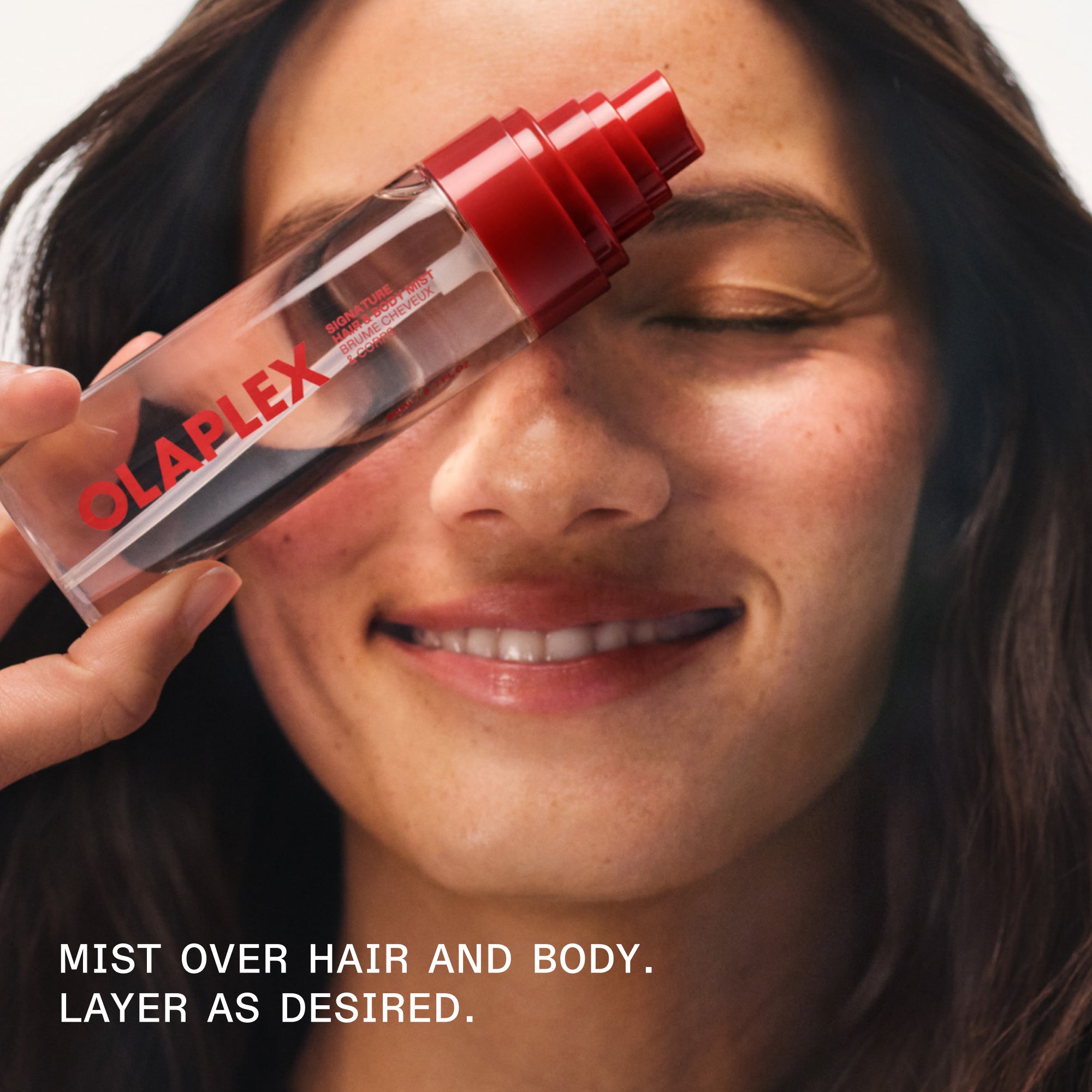 A smiling person holding an OLAPLEX hair and body mist bottle. Text overlay reads: "MIST OVER HAIR AND BODY. LAYER AS DESIRED.