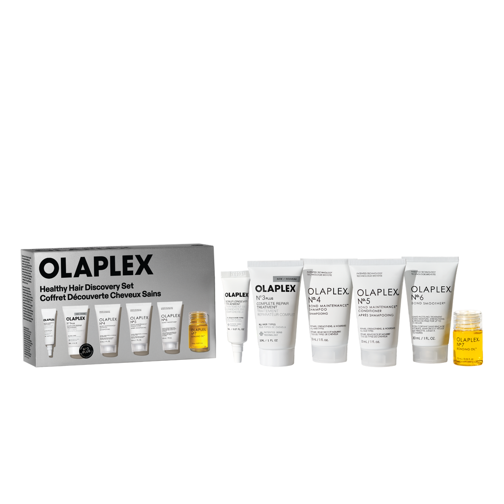 Una imagen tipo packshot retail del set Olaplex Healthy Hair Discovery sobre un fondo de tablero de ajedrez transparente. A la izquierda está la caja del producto y, a la derecha, dispuestos de forma ordenada, hay cinco tubos de tamaño viaje de color blanco (Nº.3, Nº.4, Nº.5, Nº.6 y un tubo más pequeño), junto con un pequeño frasco transparente del Nº.7 Bonding Oil con líquido amarillo en su interior. - main view