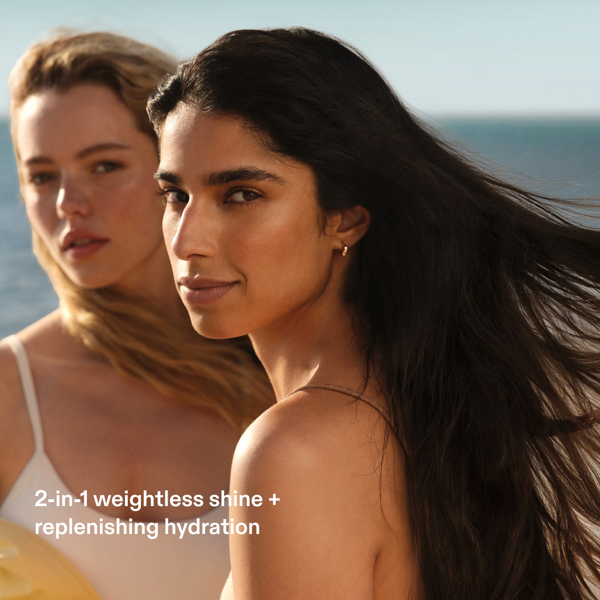 A close-up outdoor photo of two women with beautiful, healthy hair on a beach with the ocean blurred in the background. The woman in the foreground has long, dark brown hair flowing in the wind and looks toward the camera with a soft smile. White text in the lower-left corner reads: "2-in-1 weightless shine + replenishing hydration